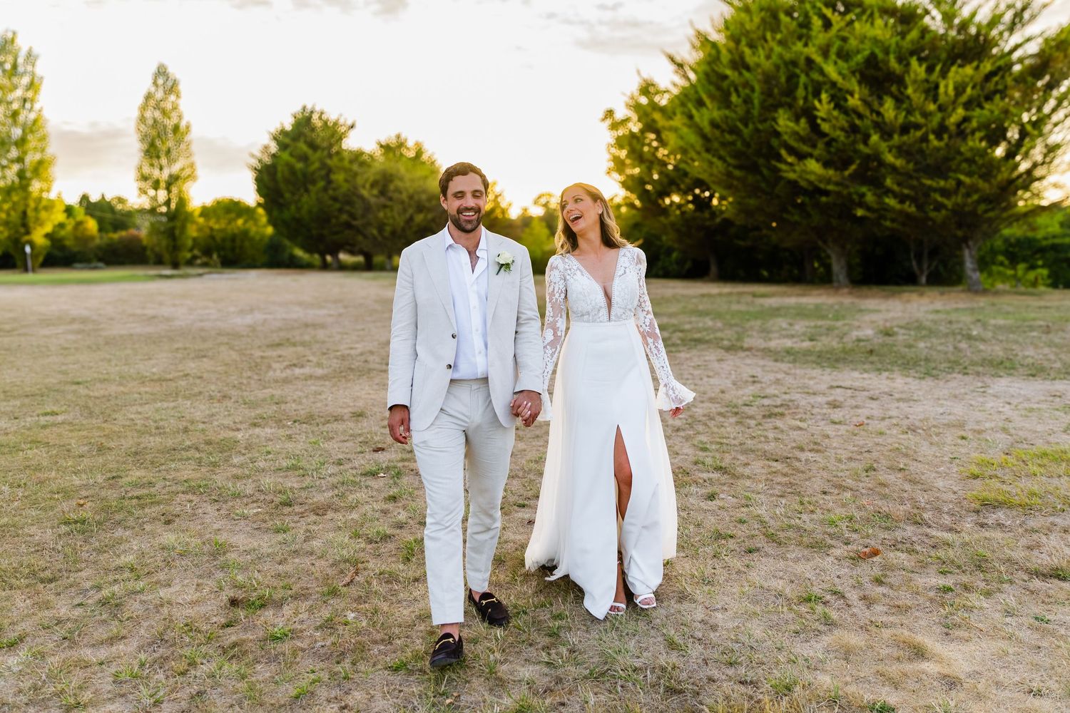 A couple in matching white outfits walking together through a grassy field at sunset.