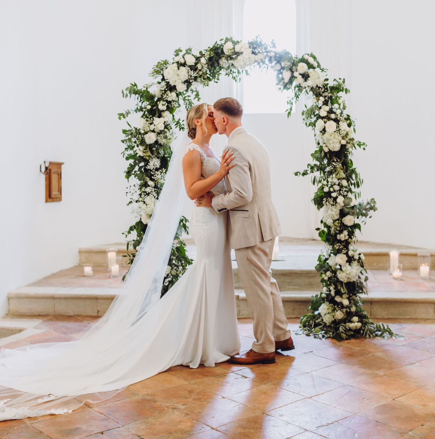 A couple shares a romantic kiss under an elegant floral arch with white flowers and greenery at their wedding ceremony.