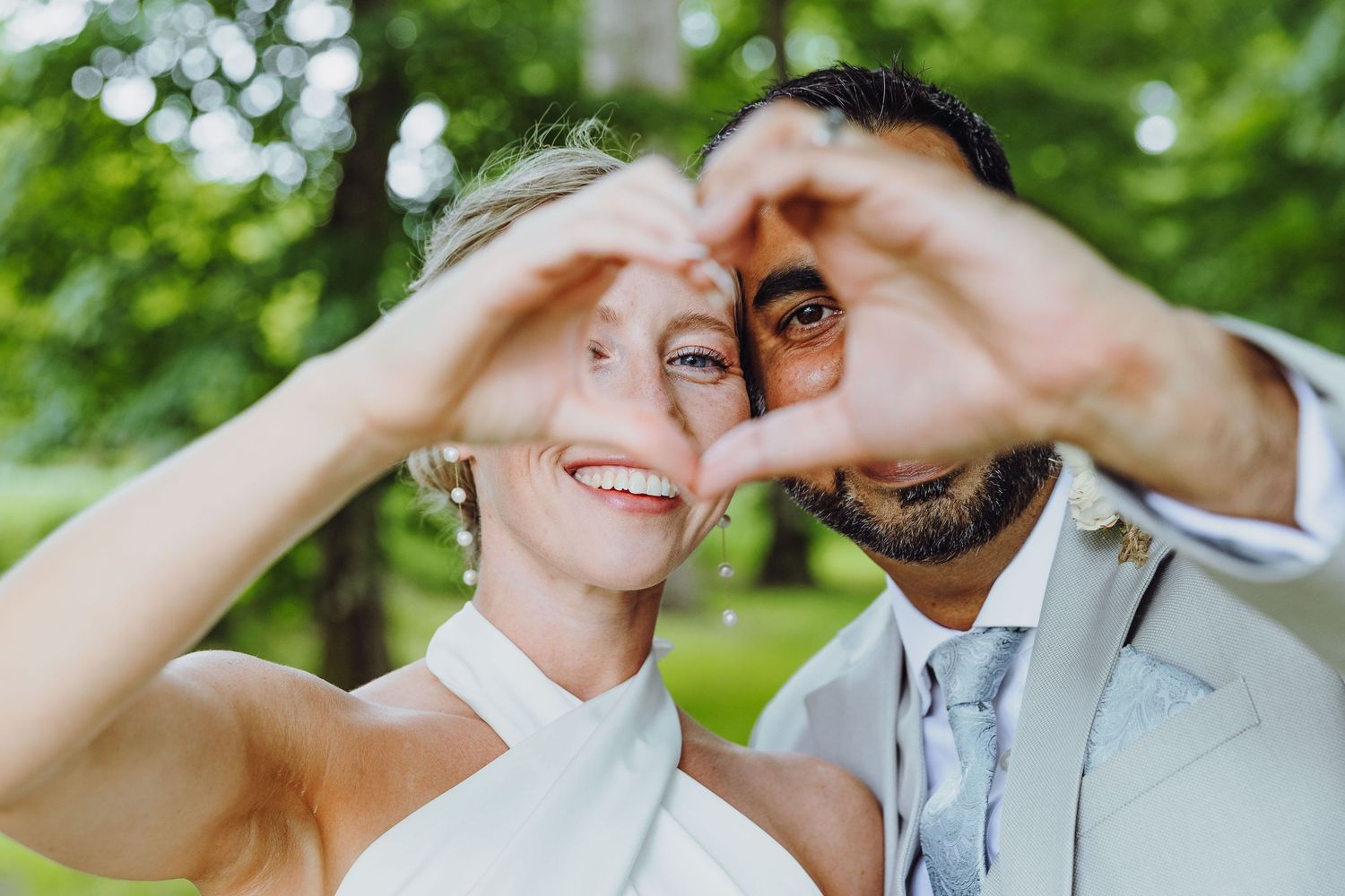 Newlywed couple making a heart shape with their hands at an outdoor wedding ceremony.