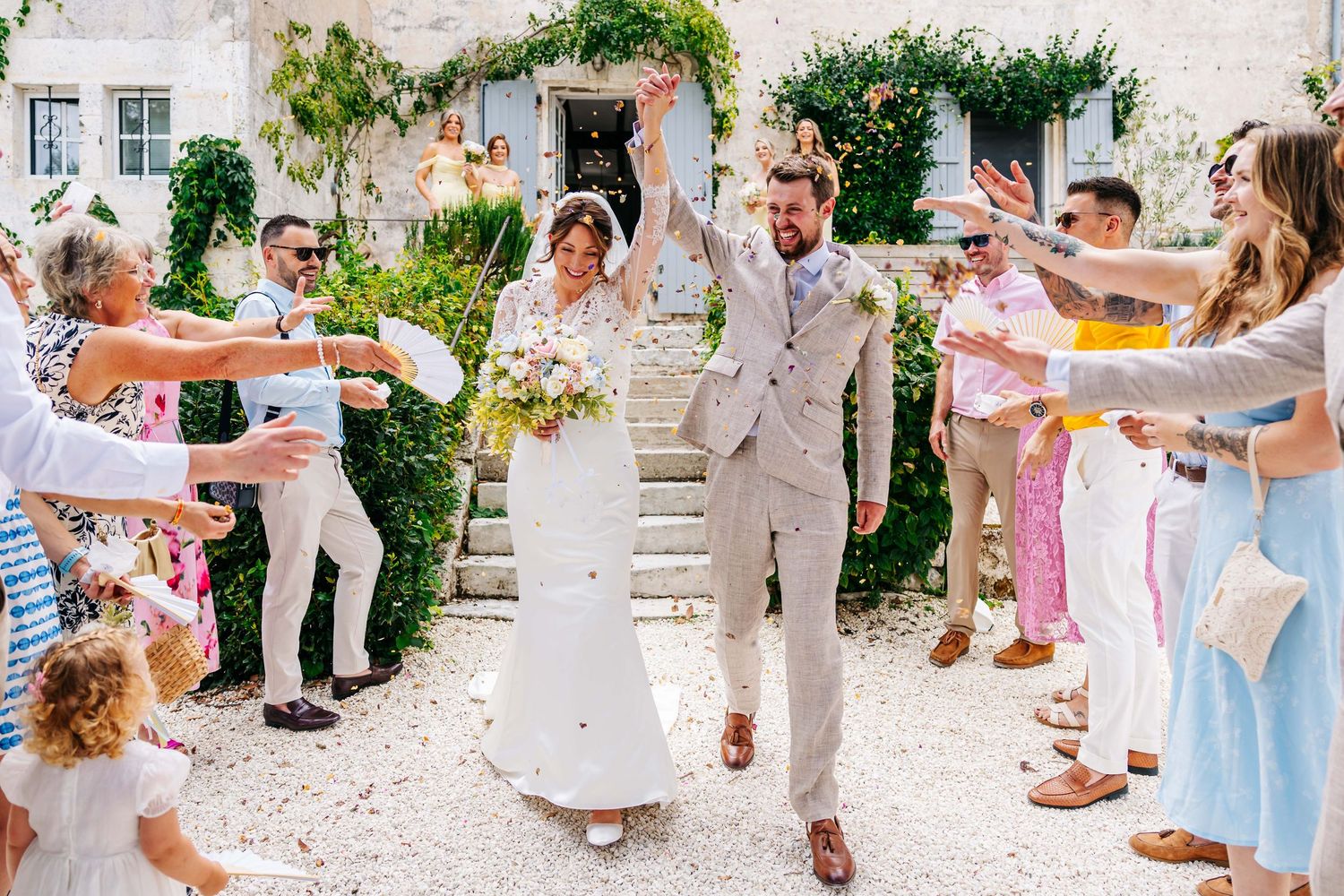 Joyful newlyweds walk through confetti shower as guests celebrate outdoor wedding at rustic European venue.