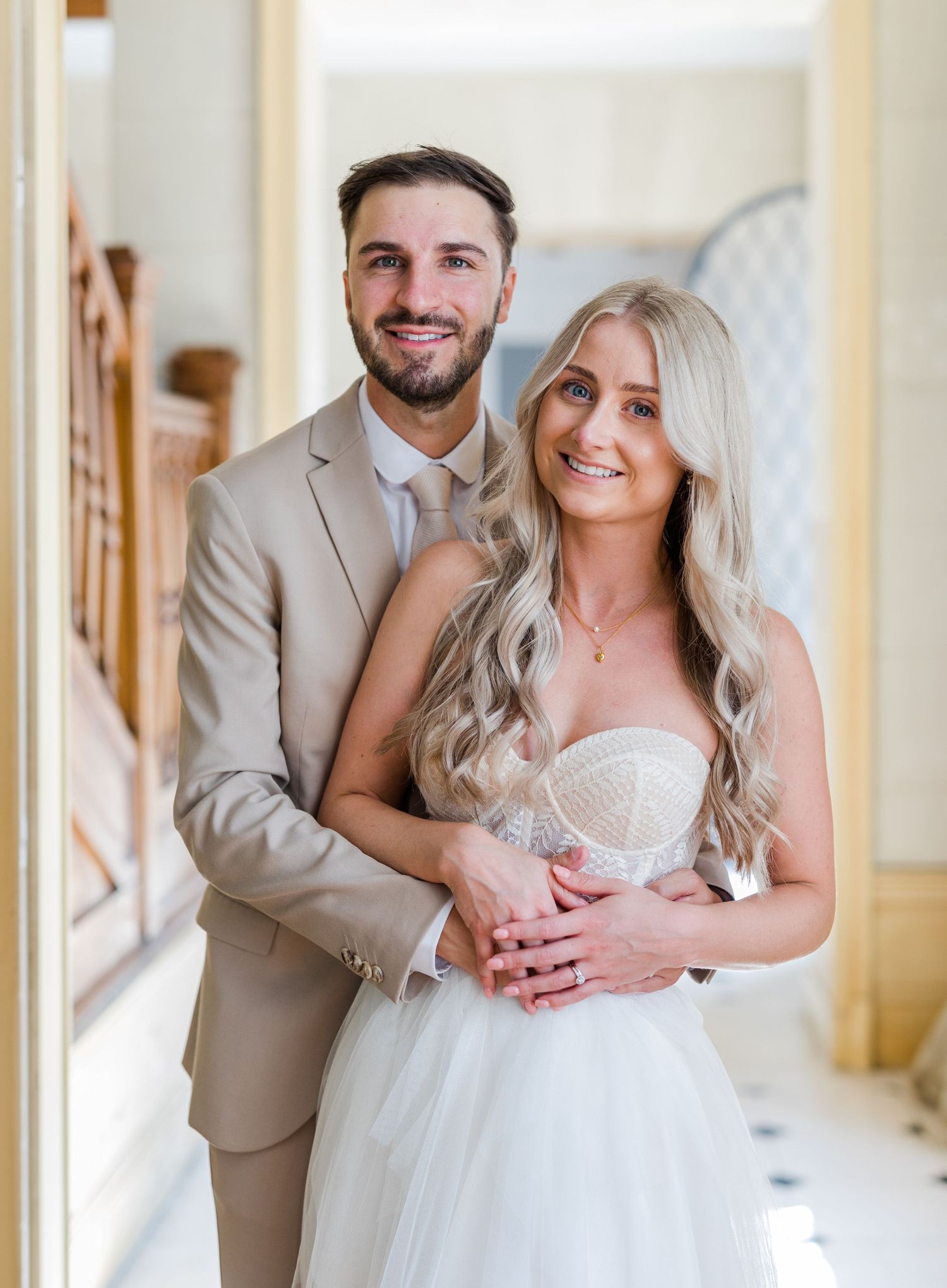 A happy couple embraces in a hallway on their wedding day, with the bride in white and groom in a beige suit.