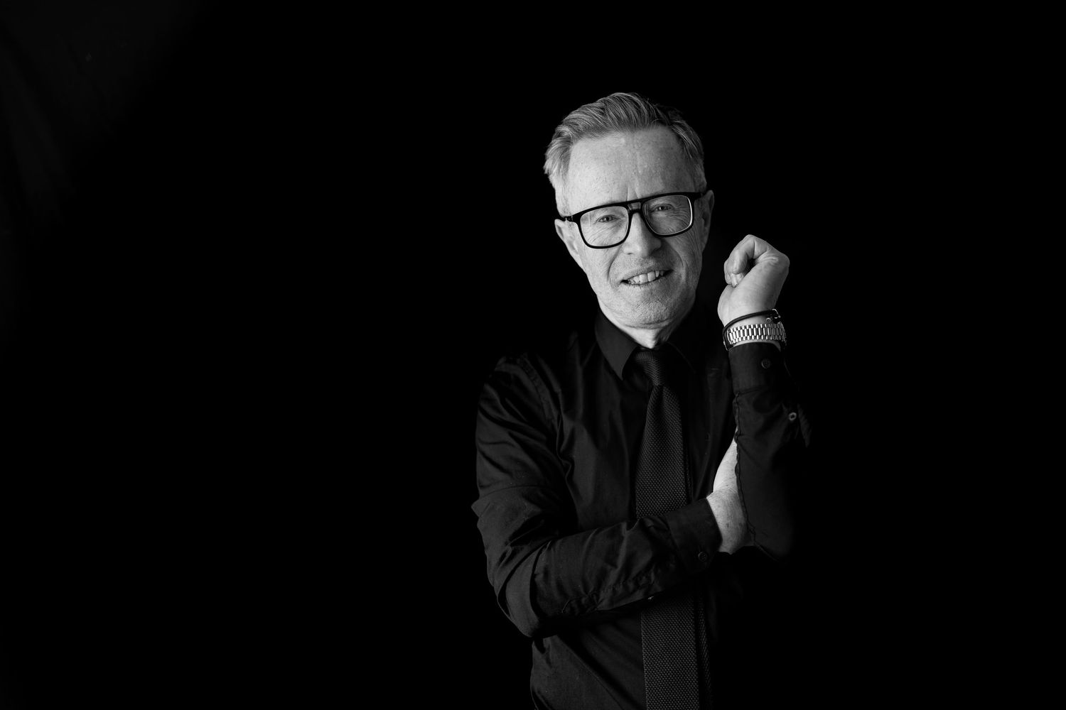 Black and white portrait of a smiling middle-aged man in a black shirt wearing glasses, posing confidently.