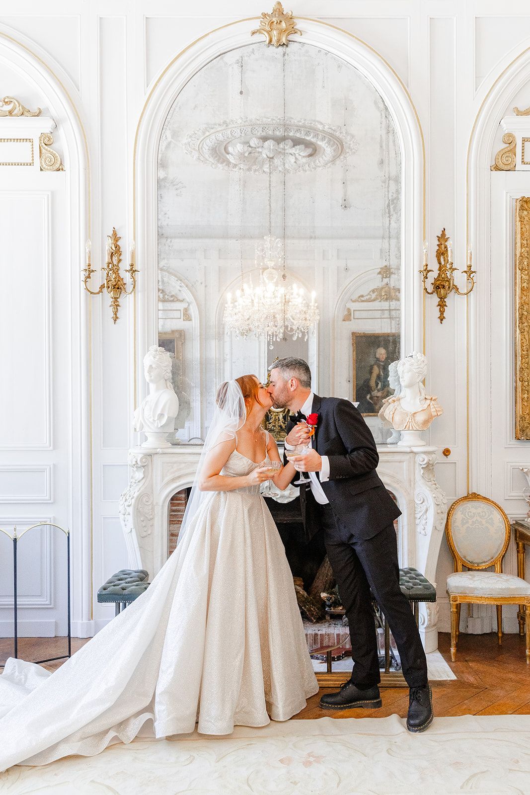 Elegant wedding couple shares champagne in ornate white ballroom with crystal chandelier and gold accents.