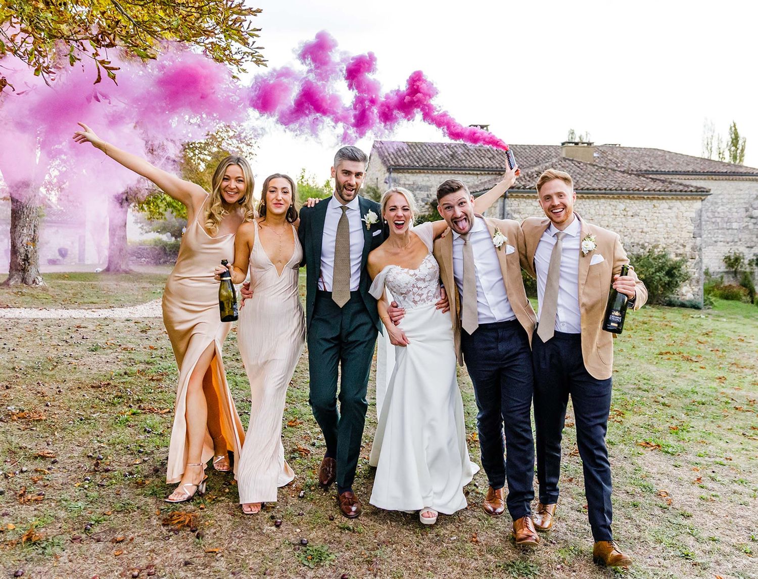 Wedding party celebrates outdoors with pink smoke bombs creating vibrant atmosphere against rustic building backdrop.