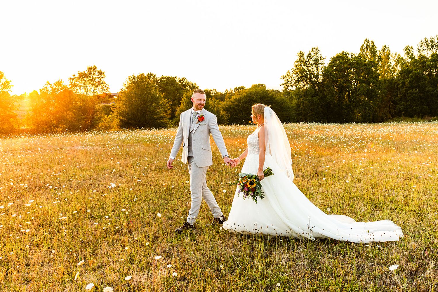 A couple in wedding attire walking hand in hand through a golden field at sunset, with a trailing white veil and bouquet.