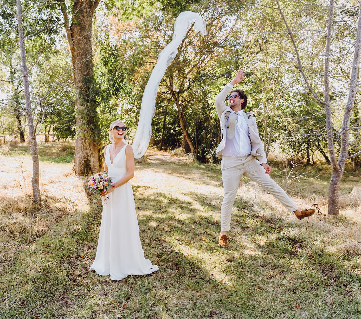 A couple poses playfully in a sun-dappled forest setting with the groom jumping in the air while the bride holds a bouquet.