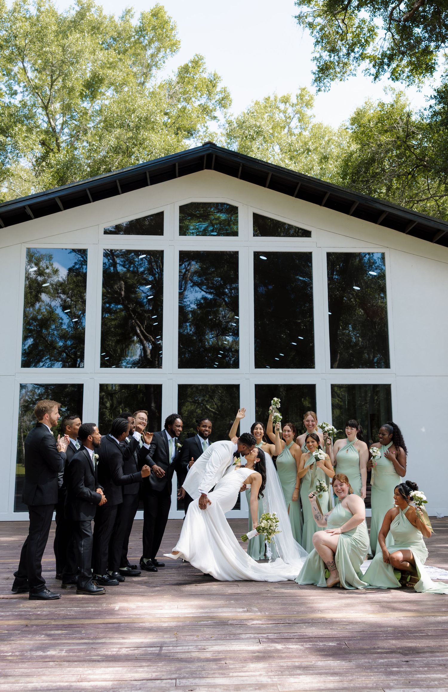 Wedding party poses outside white chapel, bride and groom share a dip kiss, groomsmen in black suits, bridesmaids in sage green gowns.