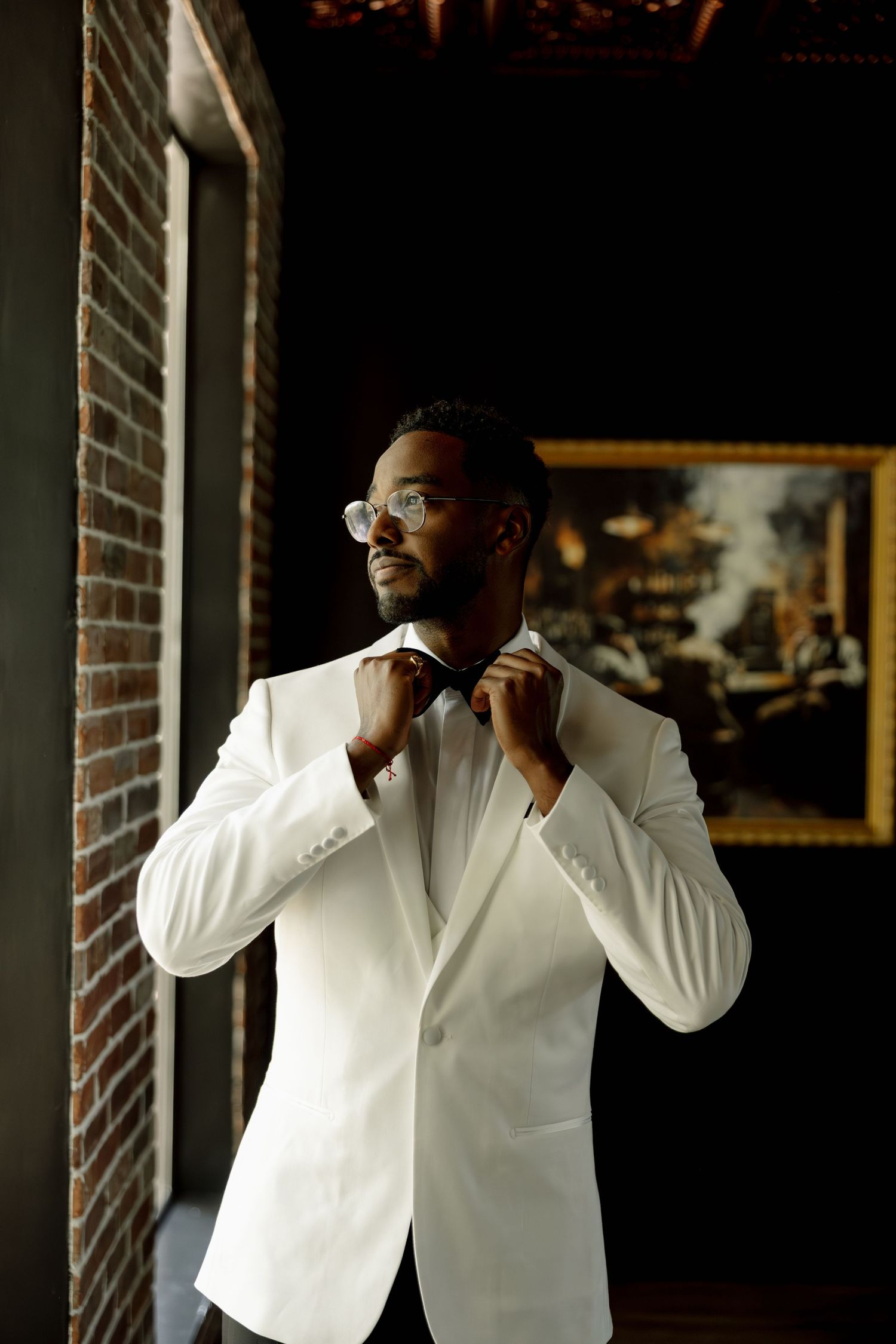 A well-dressed Black man in a white tuxedo jacket adjusts his bow tie while standing near a brick wall.