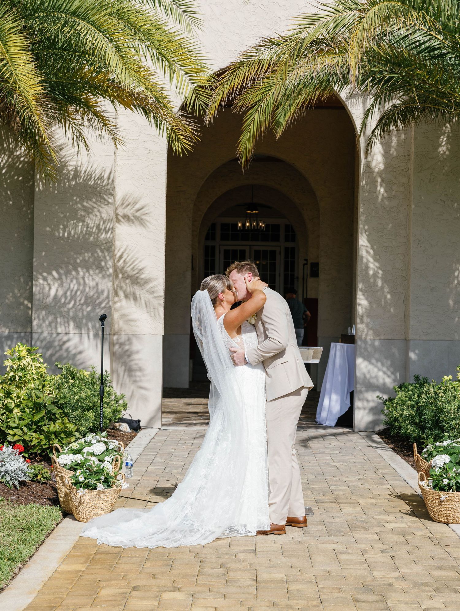 Bride and groom share first kiss outside elegant venue entrance with palm trees and floral arrangements on sunny wedding day.