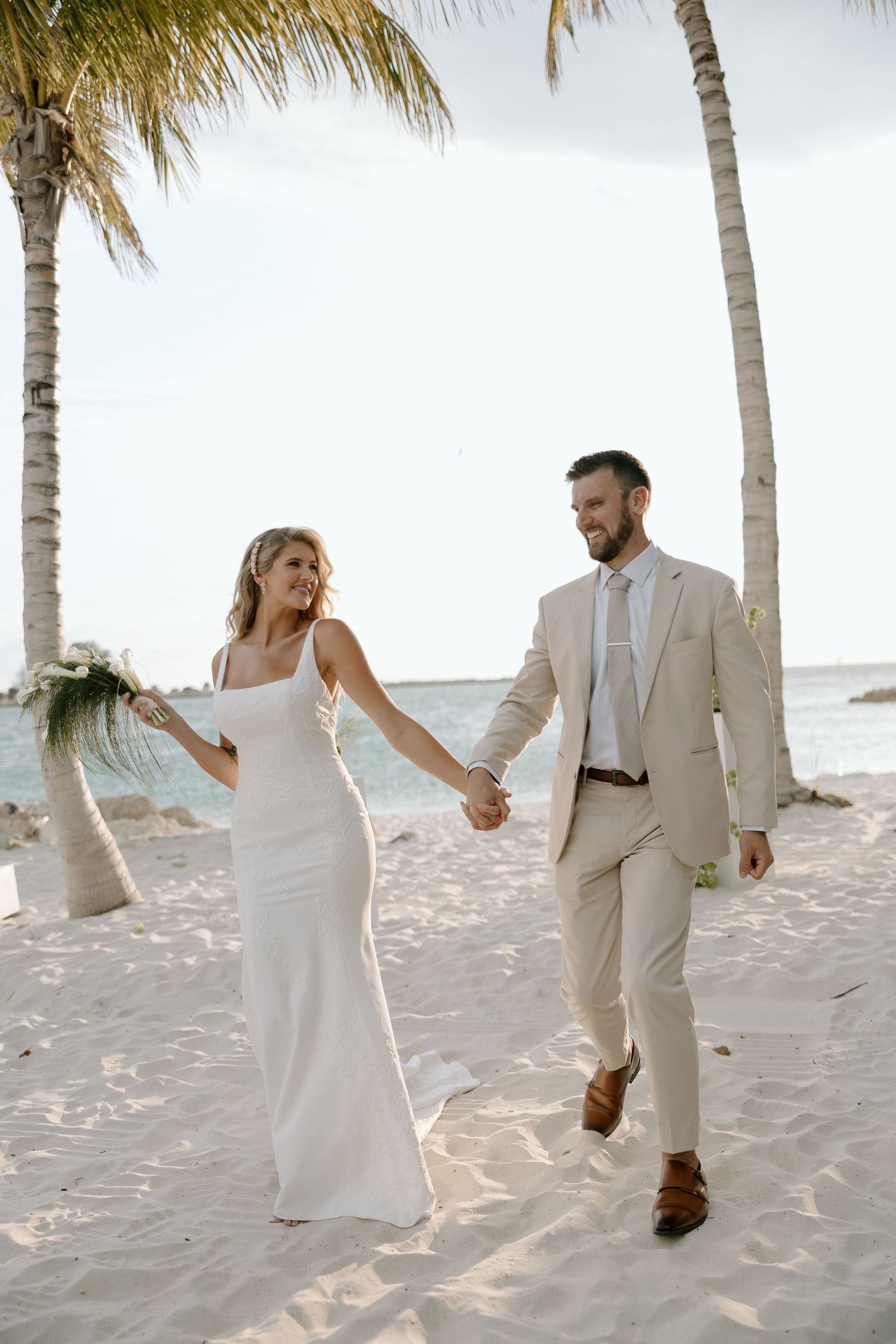 Bride and groom walk hand-in-hand on a tropical beach, she in a white gown, he in a beige suit.