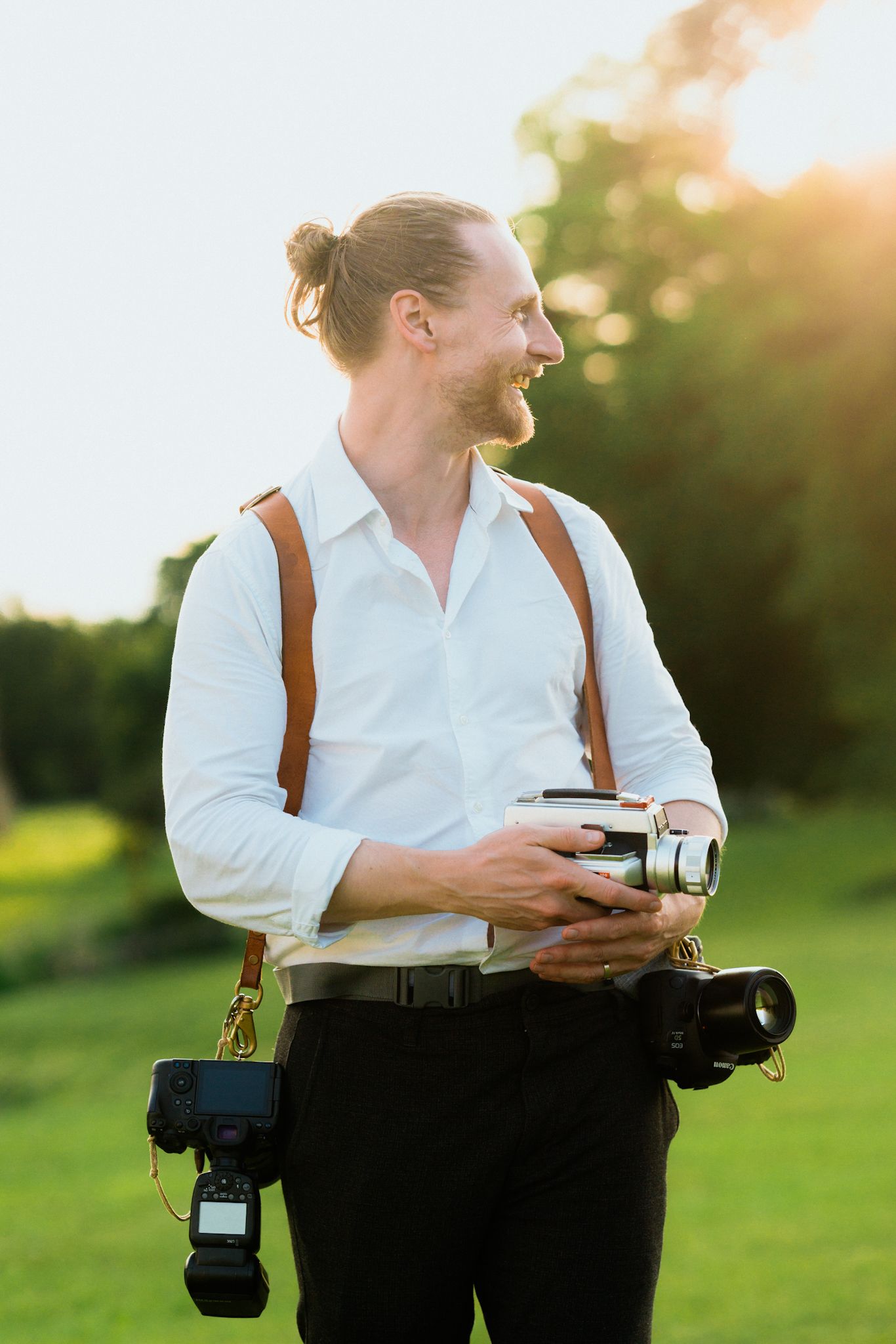 Person in white shirt and dark pants holds camera while walking in sunlit grassy field at golden hour.