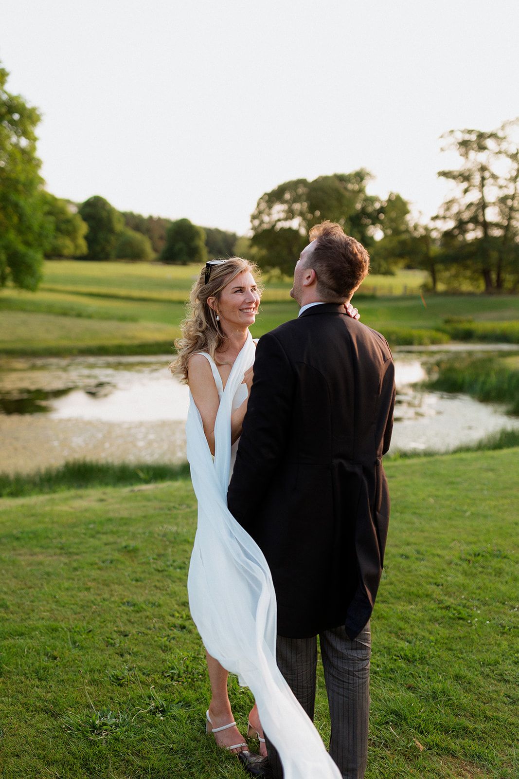 A bride and groom share a romantic moment on a lush green lawn with a pond and trees in the background at sunset.