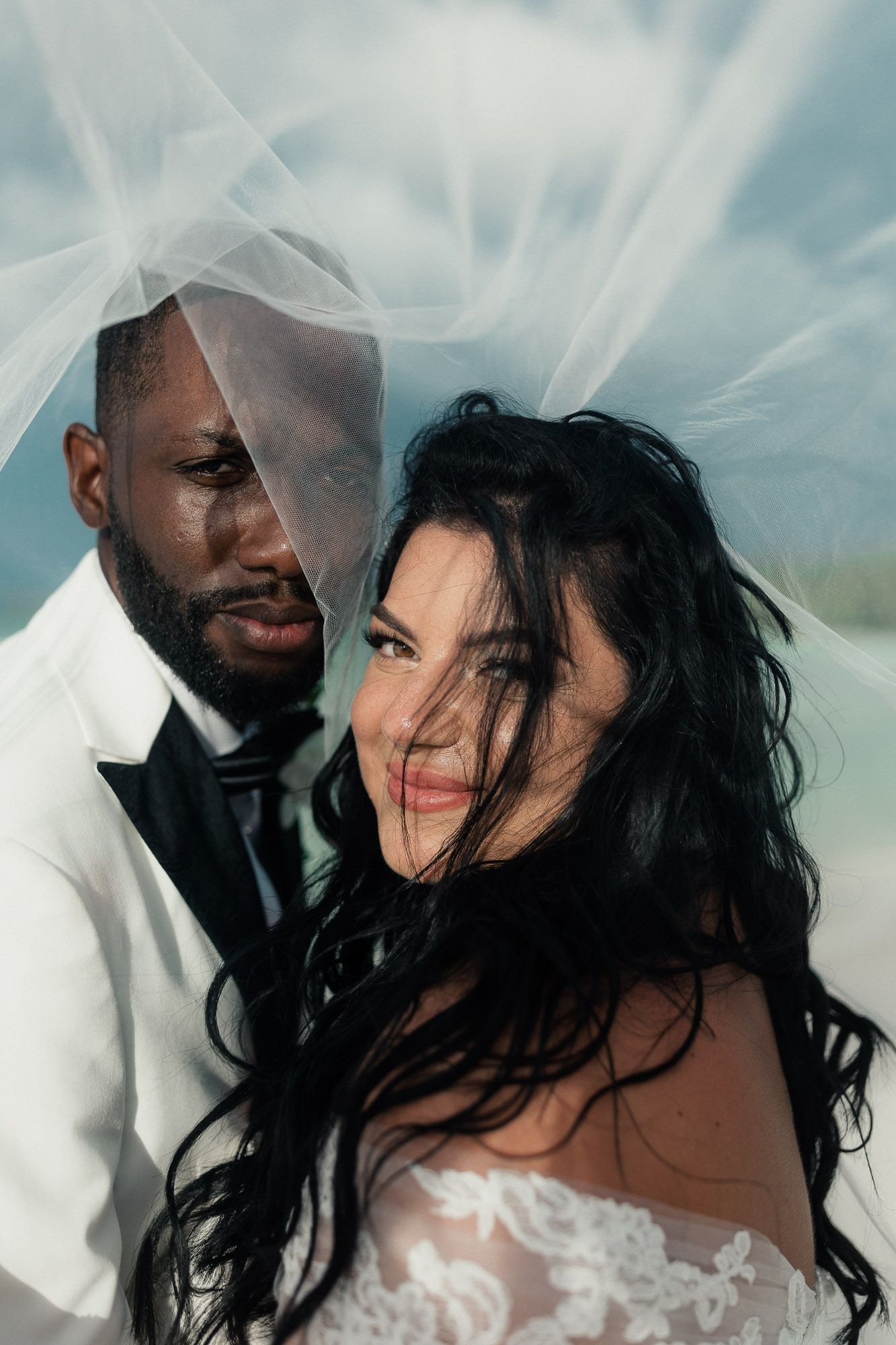 A romantic wedding couple embraces under a cloudy sky with flowing white veil creating a dreamy atmosphere.