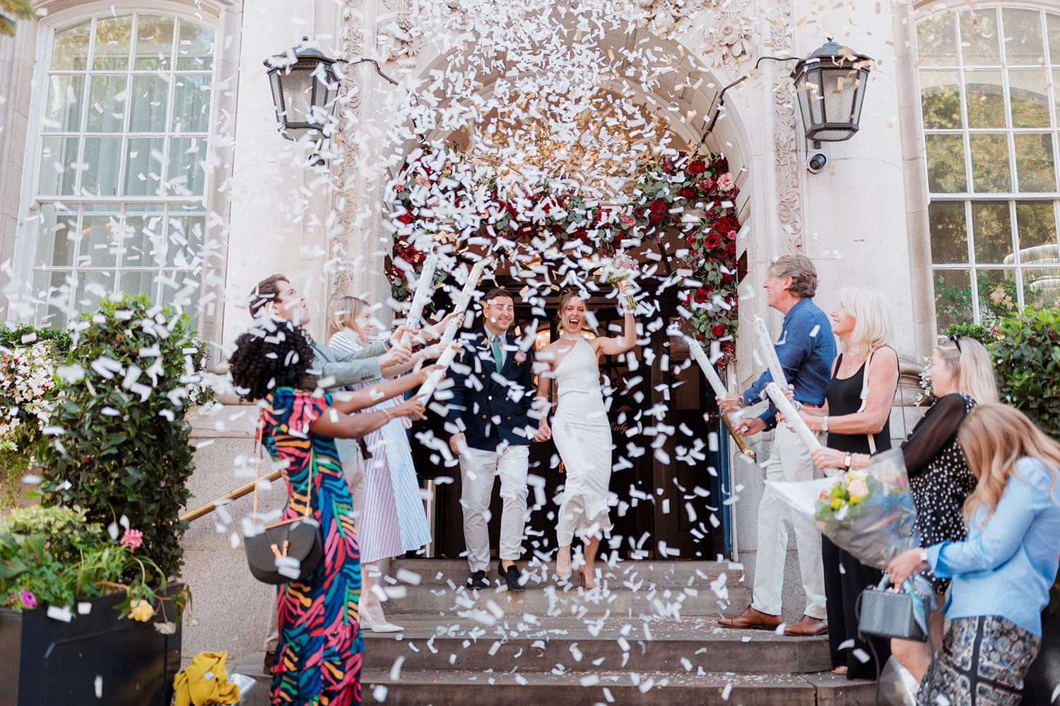 Wedding guests celebrate with confetti during an outdoor ceremony at an elegant venue with white columns and windows.