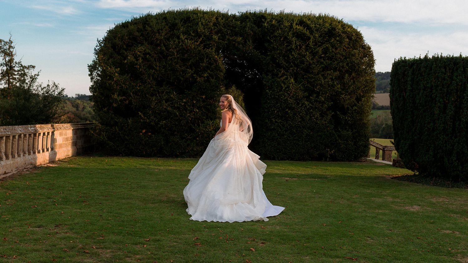 A bride in a flowing white gown poses on a lush green lawn with trees and a historic building in the background.