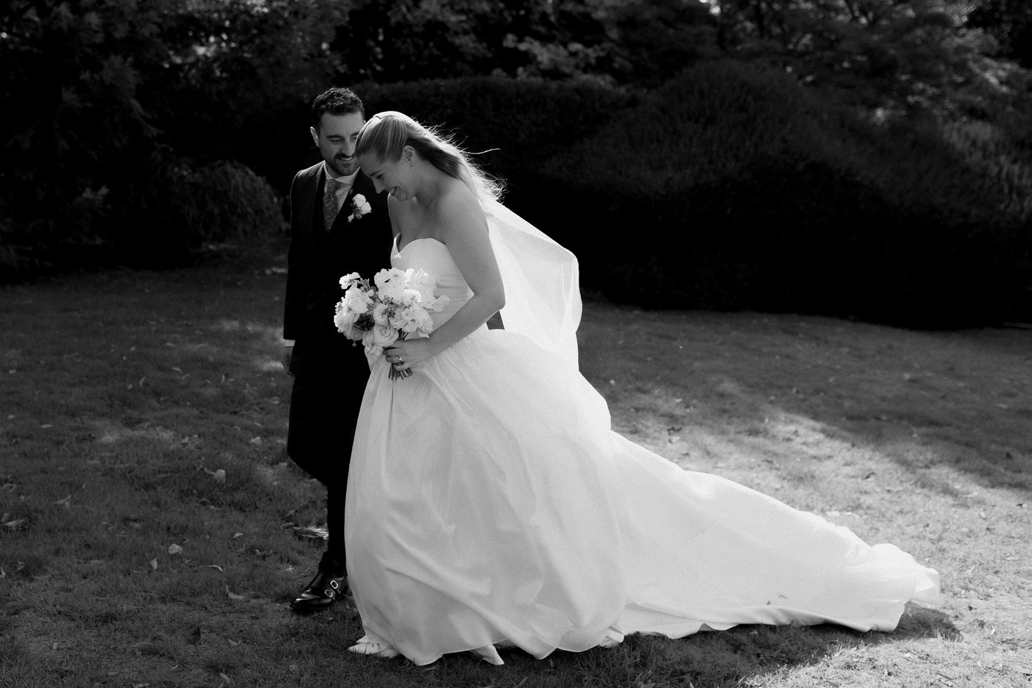 A black and white wedding photograph showing a couple embracing outdoors in a garden setting.