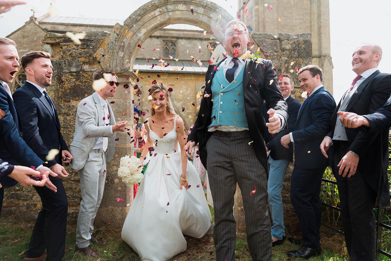 Wedding guests gather outside a historic stone building while throwing rose petals during a joyful celebration.