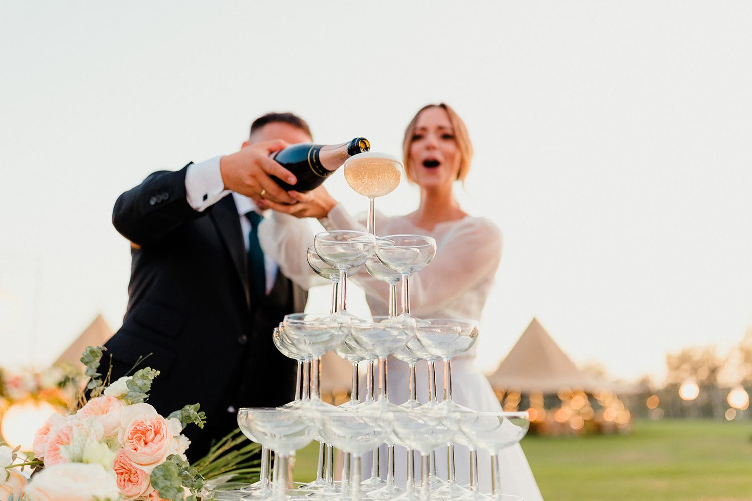A champagne tower being poured at an elegant outdoor wedding reception during sunset.