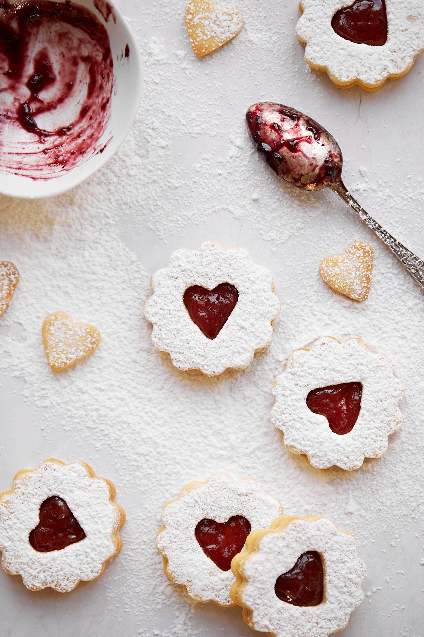 Homemade Linzer cookies with heart cutouts and jam filling, displayed with a used spoon and bowl.