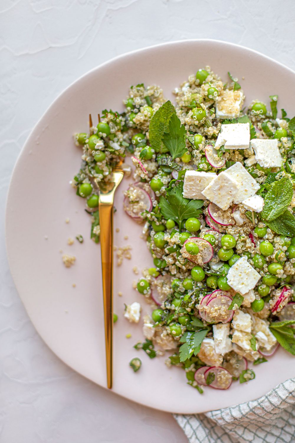 Vibrant quinoa salad with feta, peas and radishes on a white plate with gold utensils.