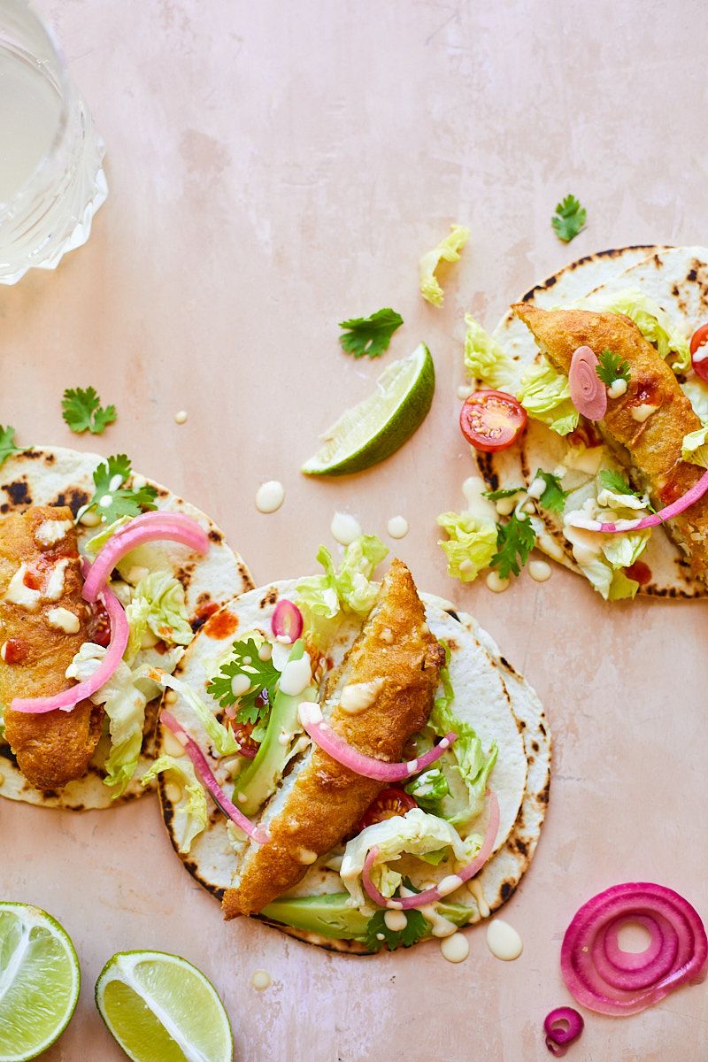 Fish tacos garnished with fresh herbs, pickled onions and lime on a light pink background.