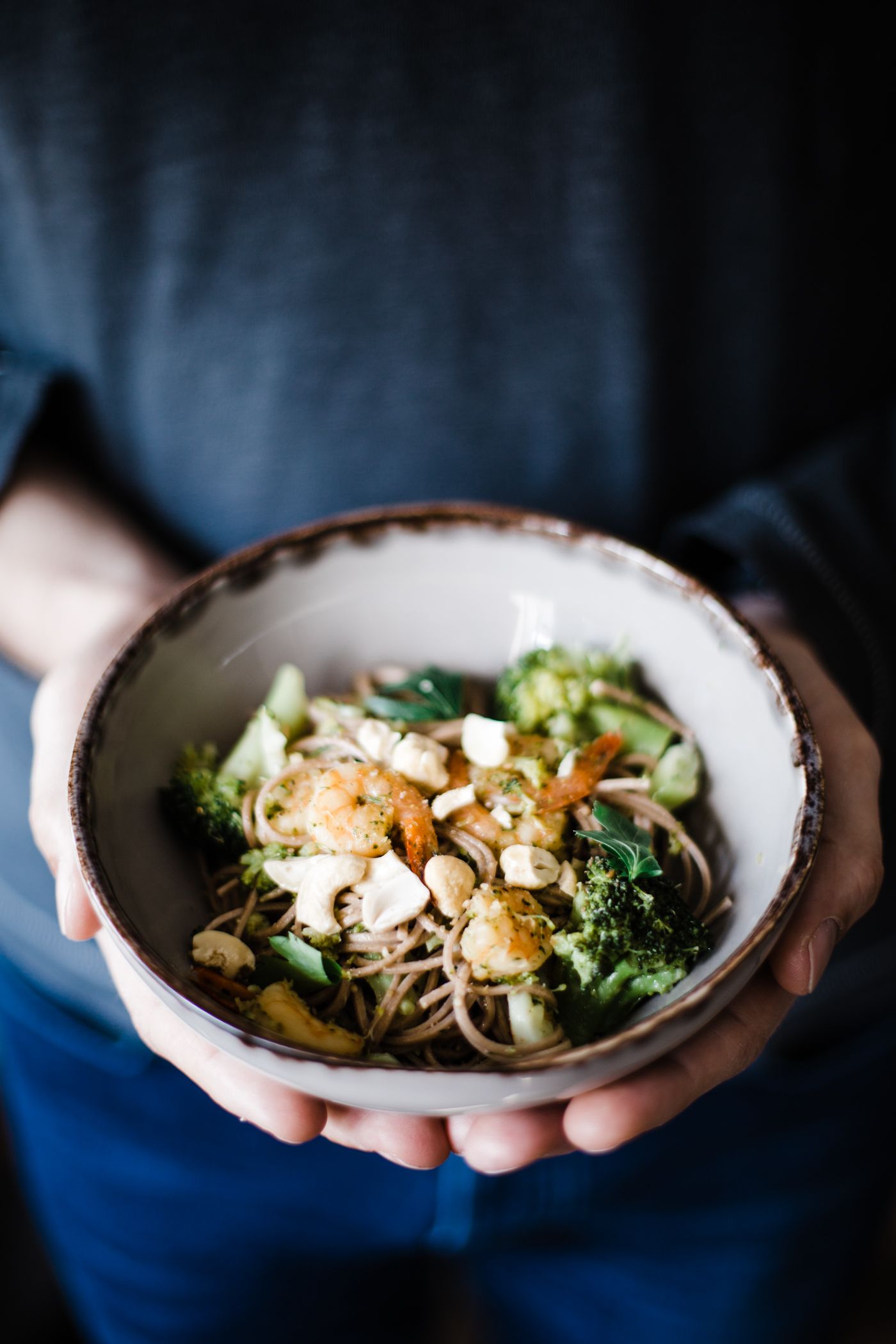 Noodle bowl with broccoli, mushrooms and almonds held in hands against dark background.
