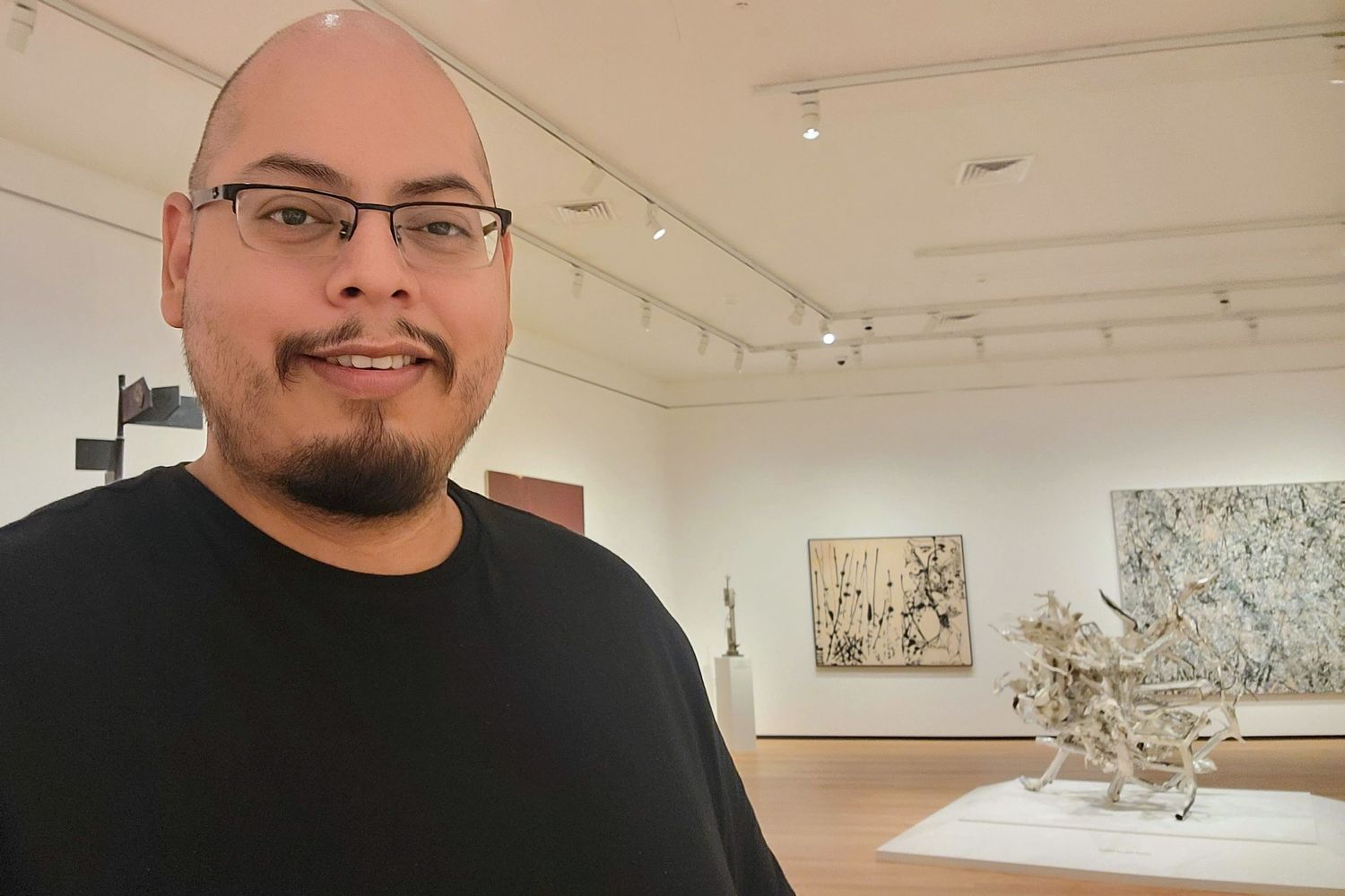 A person wearing a black shirt stands in a white-walled art gallery with framed artwork and sculptures on display.