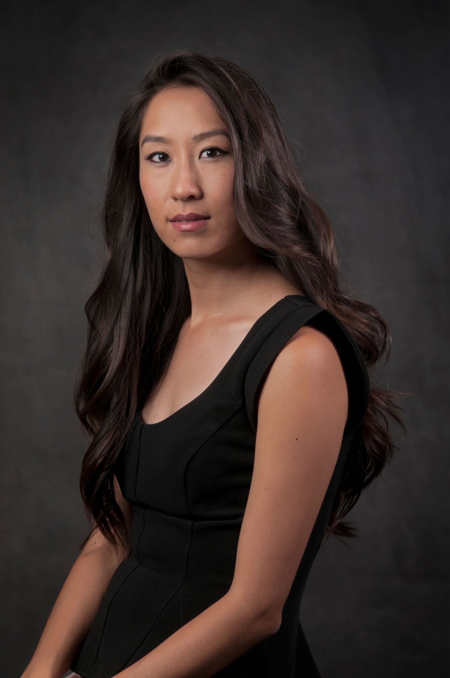 Professional studio portrait against dark backdrop shows person with long dark wavy hair wearing a sleek black sleeveless dress.