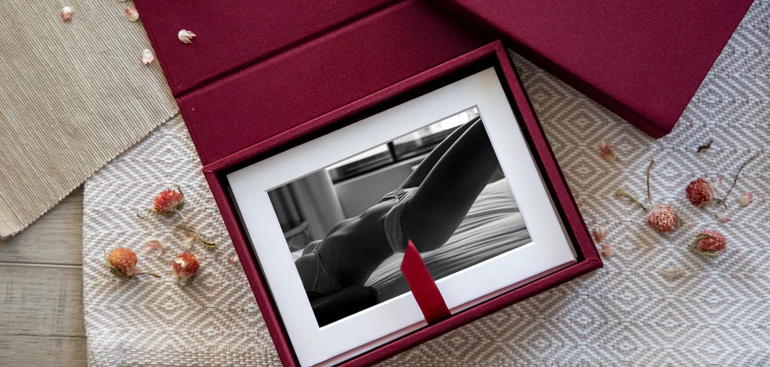 A framed black and white photograph in a burgundy gift box with red rose petals scattered on a wooden surface.