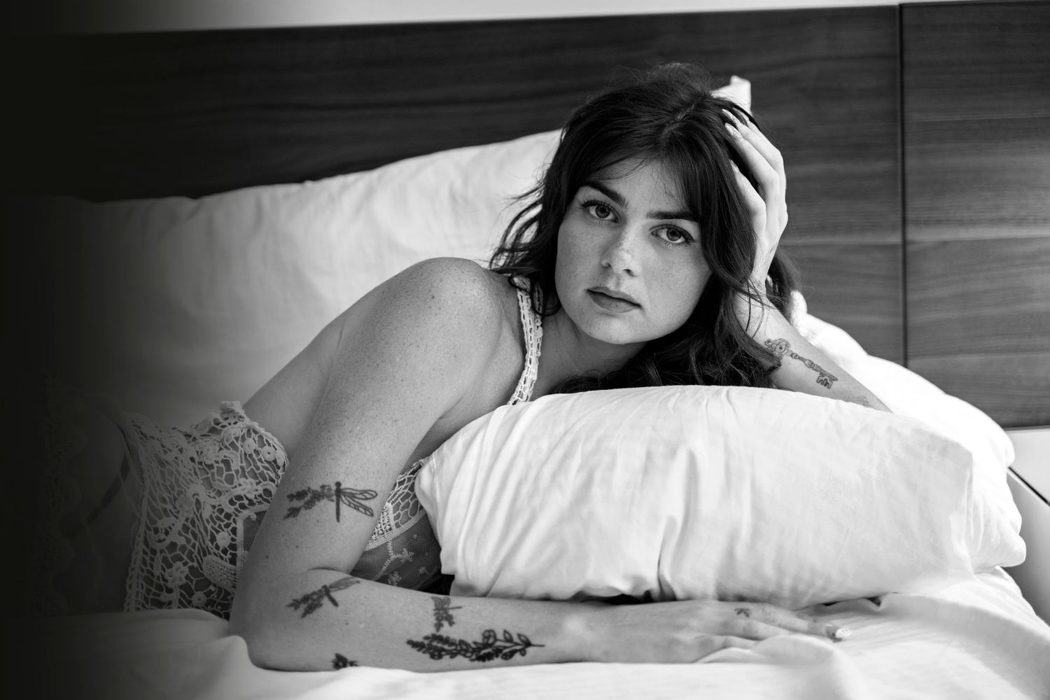 Black and white photo of a tattooed woman lying on a bed with a pillow, gazing thoughtfully at the camera.