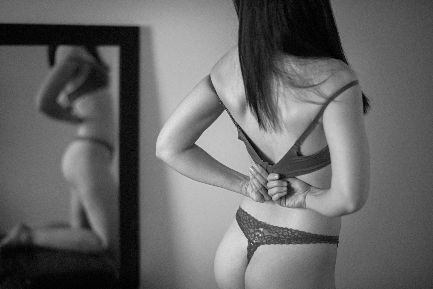Black and white photograph showing a figure adjusting clothes in front of a mirror in an intimate setting.