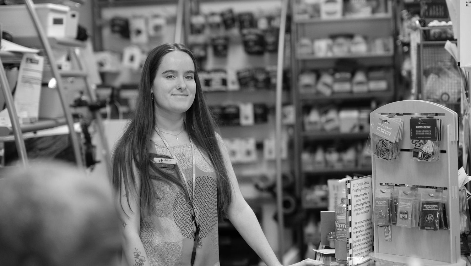 Woman shopping in pharmacy aisle examines product label while wearing casual attire and surgical mask.