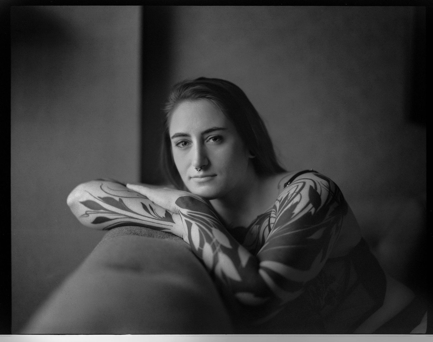 A black and white portrait photograph in dramatic lighting shows a figure resting their head on folded arms.