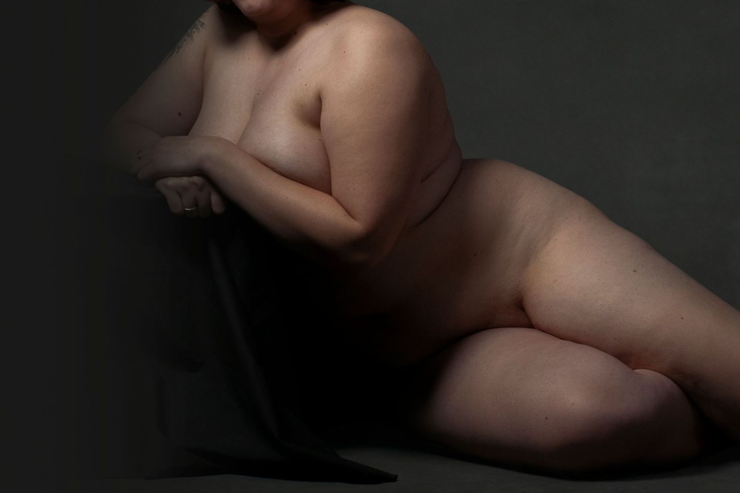Artistic nude fine art photography of a plus-size woman reclining gracefully against a dark chair in moody studio lighting.