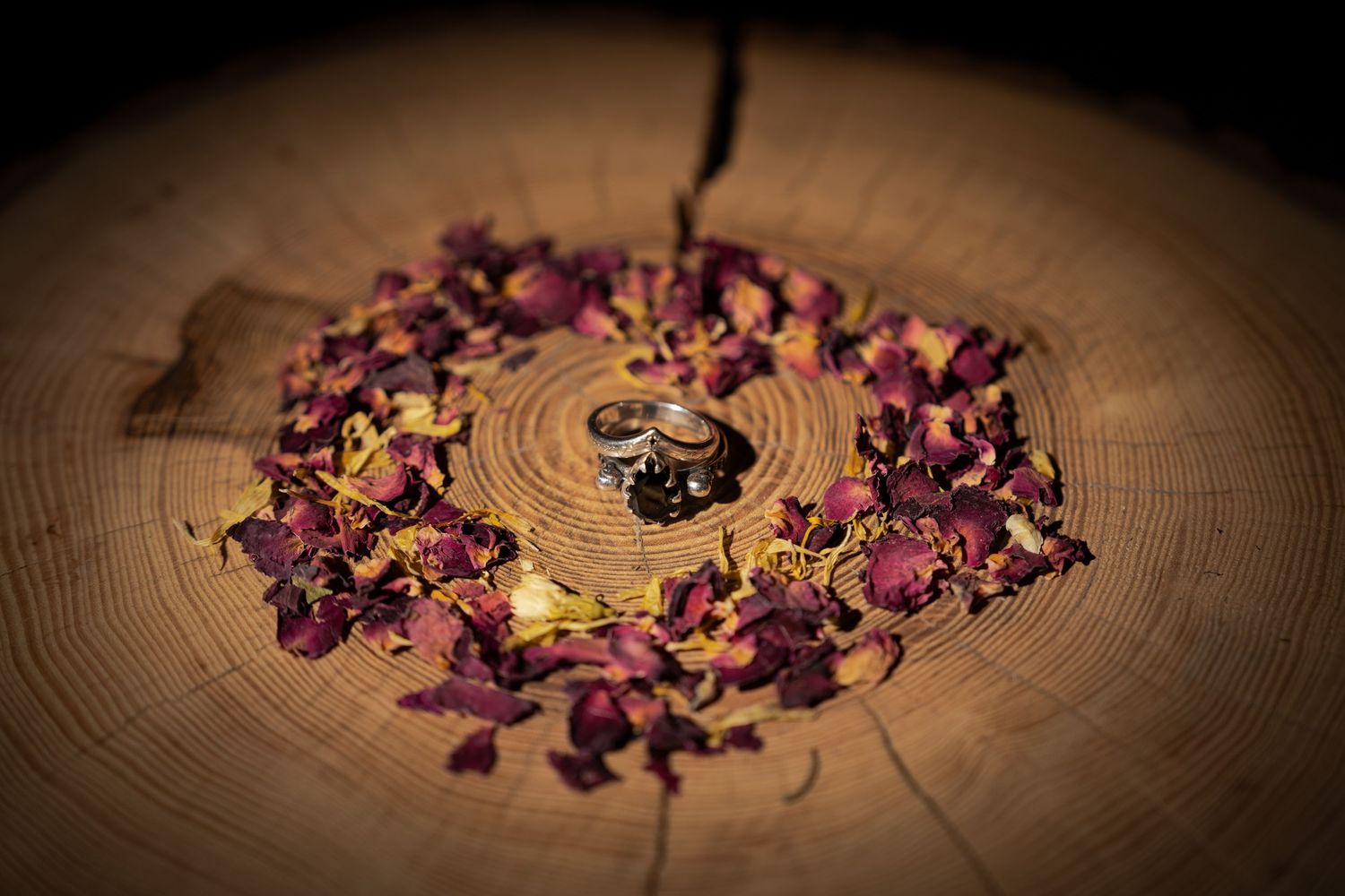 Dried rose petals arranged in a heart around a ring on a wooden tree stump.