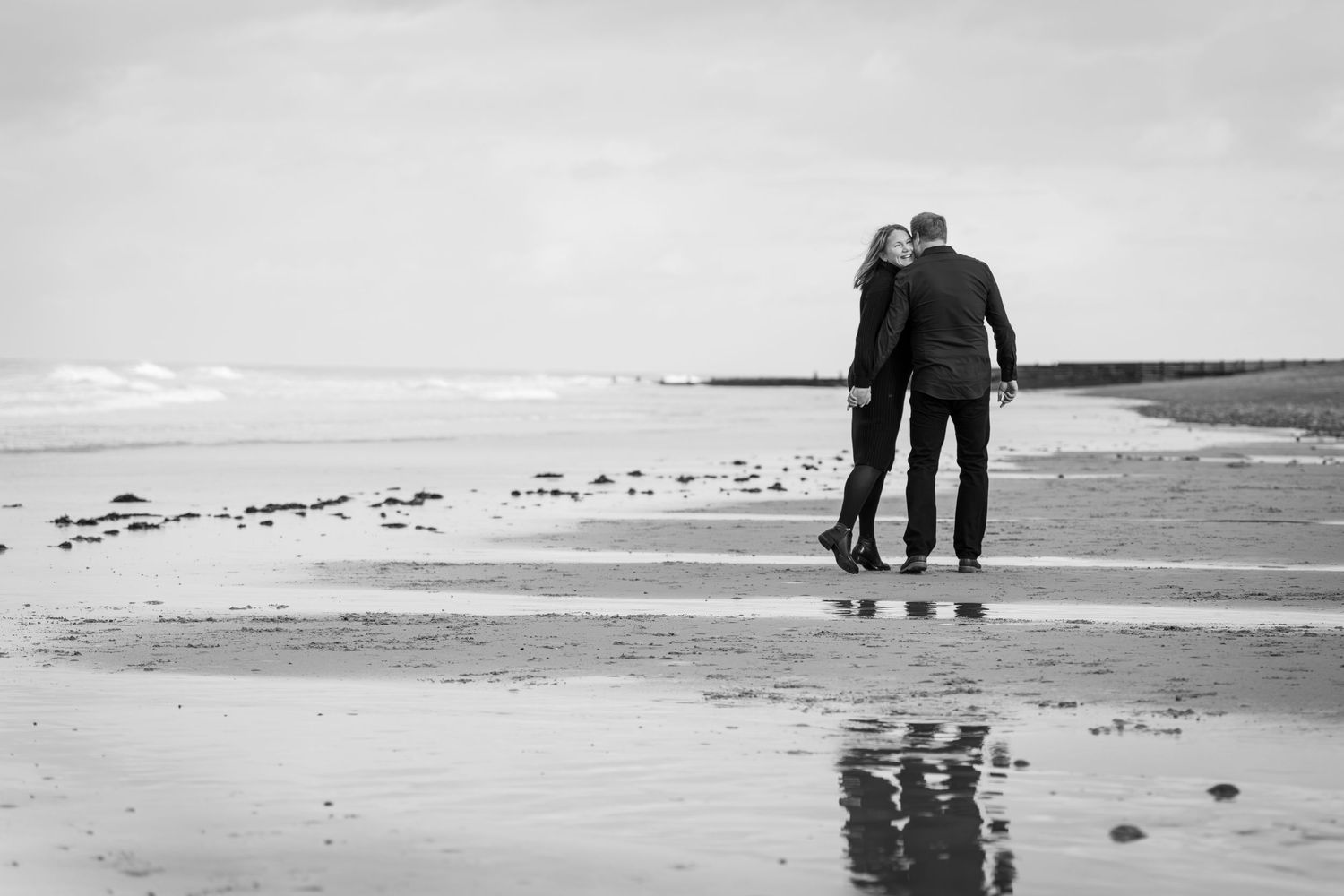 Black and white photo of a couple standing together on a wet beach, gazing at the ocean waves.