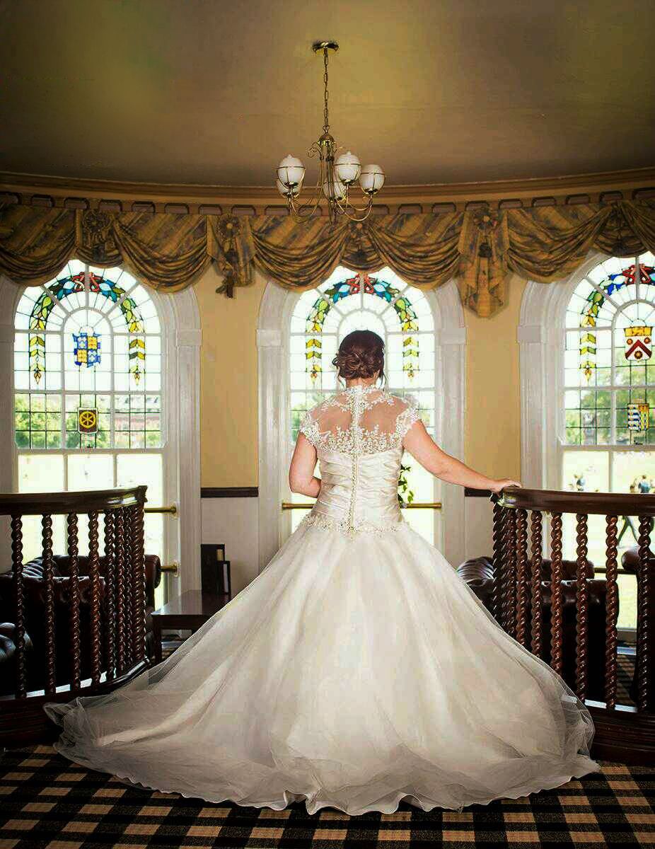 Bride in a white lace-back wedding gown with a long flowing train stands on a balcony facing stained glass windows and gold drapes.