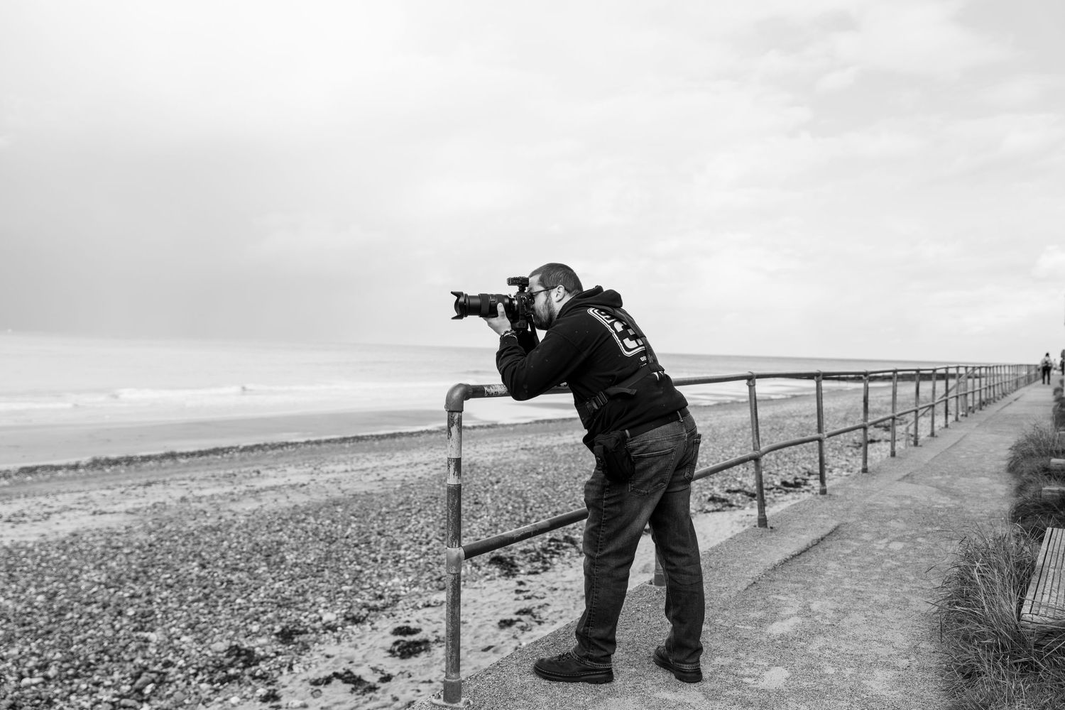 Photographer leaning on seaside railing capturing ocean views in black and white coastal landscape.