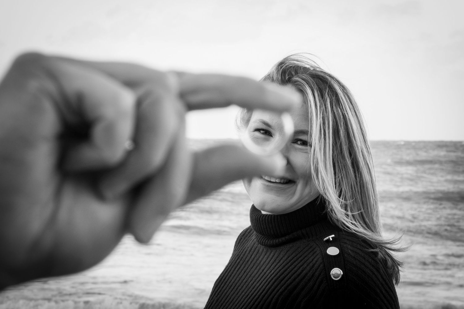 Black and white photo of smiling blonde woman framed by hand fingers near ocean beach.