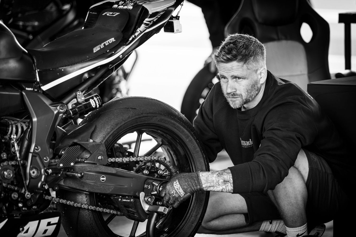 Tattooed mechanic crouches beside a racing motorcycle in black and white, adjusting the rear wheel and chain in a pit garage.