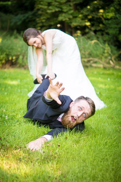 Playful bride in a white wedding dress drags a groom in a navy suit by the leg across a green lawn as he reaches out dramatically.