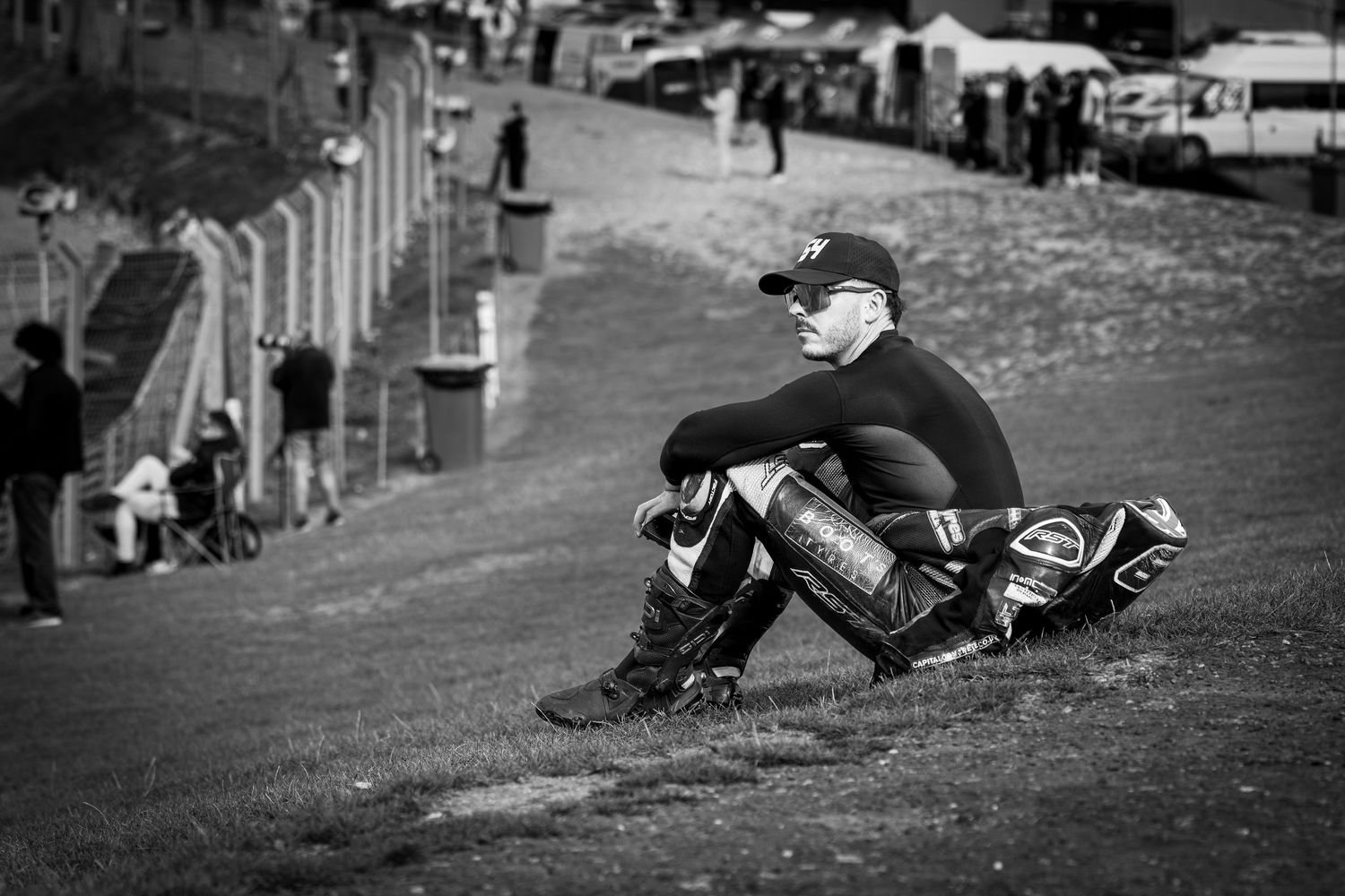 Black and white photo of a motorcycle racer in leather suit sitting on a grass hill at a racing circuit.