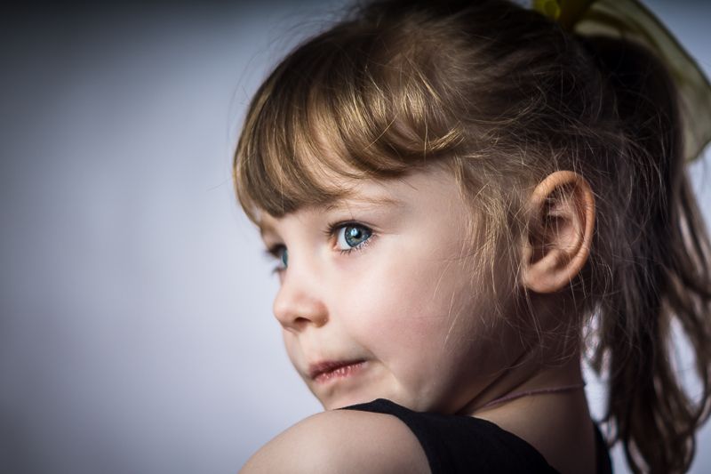 Close-up portrait of a young girl with blue eyes and light brown hair with bangs, looking to the side against a soft gray background.
