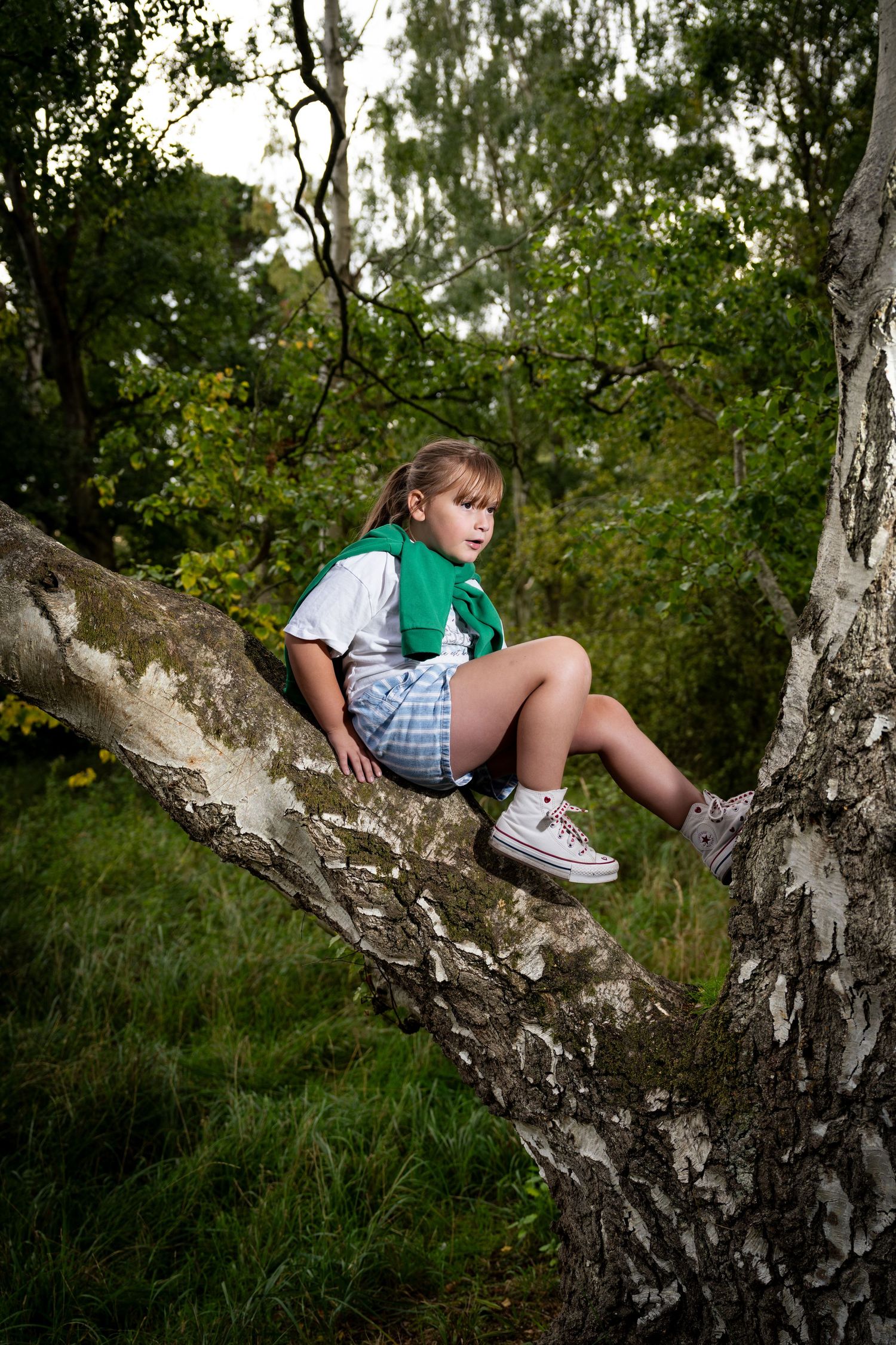 Young girl sitting on a birch tree branch in a forest wearing a green vest and plaid shorts.