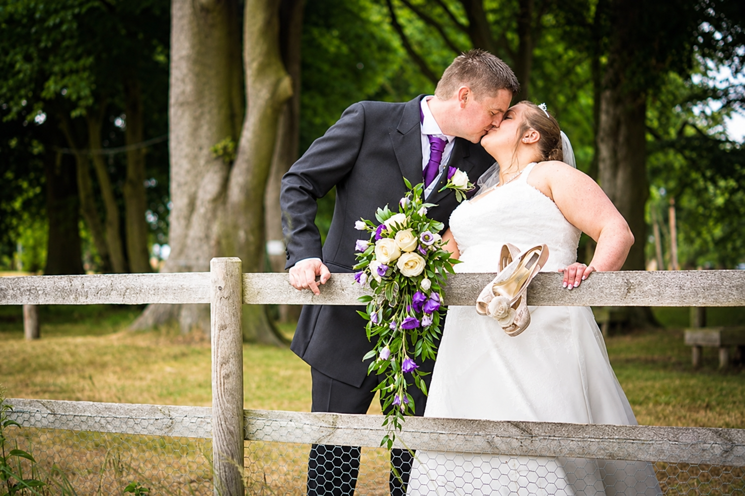 Bride and groom share a kiss beside a wooden fence in a leafy park, the bride holding a purple and cream bouquet and a straw hat.
