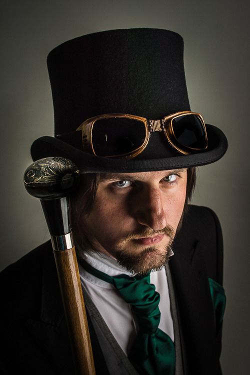 Man in steampunk attire wearing a black top hat with brass goggles, green cravat and black coat, holding an ornate wooden cane.