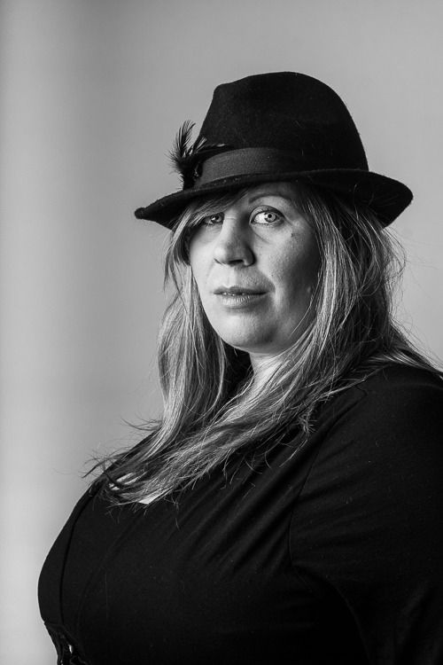 Black and white portrait of a woman with long hair wearing a dark fedora hat with a feather accent and a black top.