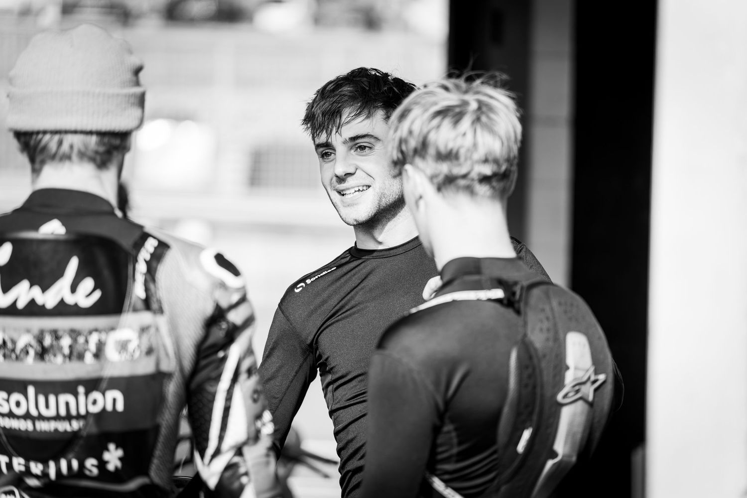 Black and white candid photo of a smiling young male racing driver chatting at a motorsport paddock.