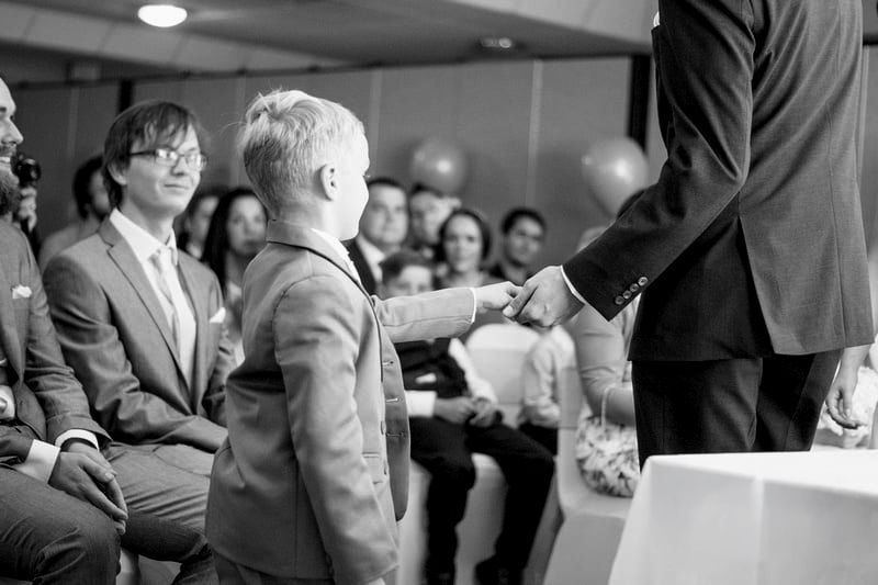 Black and white photo of a young boy in a suit shaking hands with a man as seated wedding guests look on.