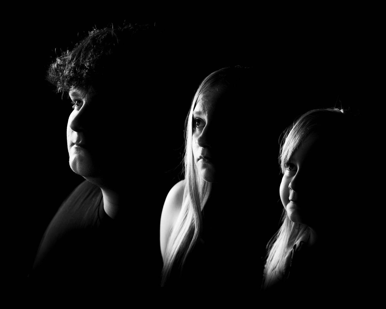 Black and white low-key profile portrait of three people in silhouette, lit from the side against a dark background.